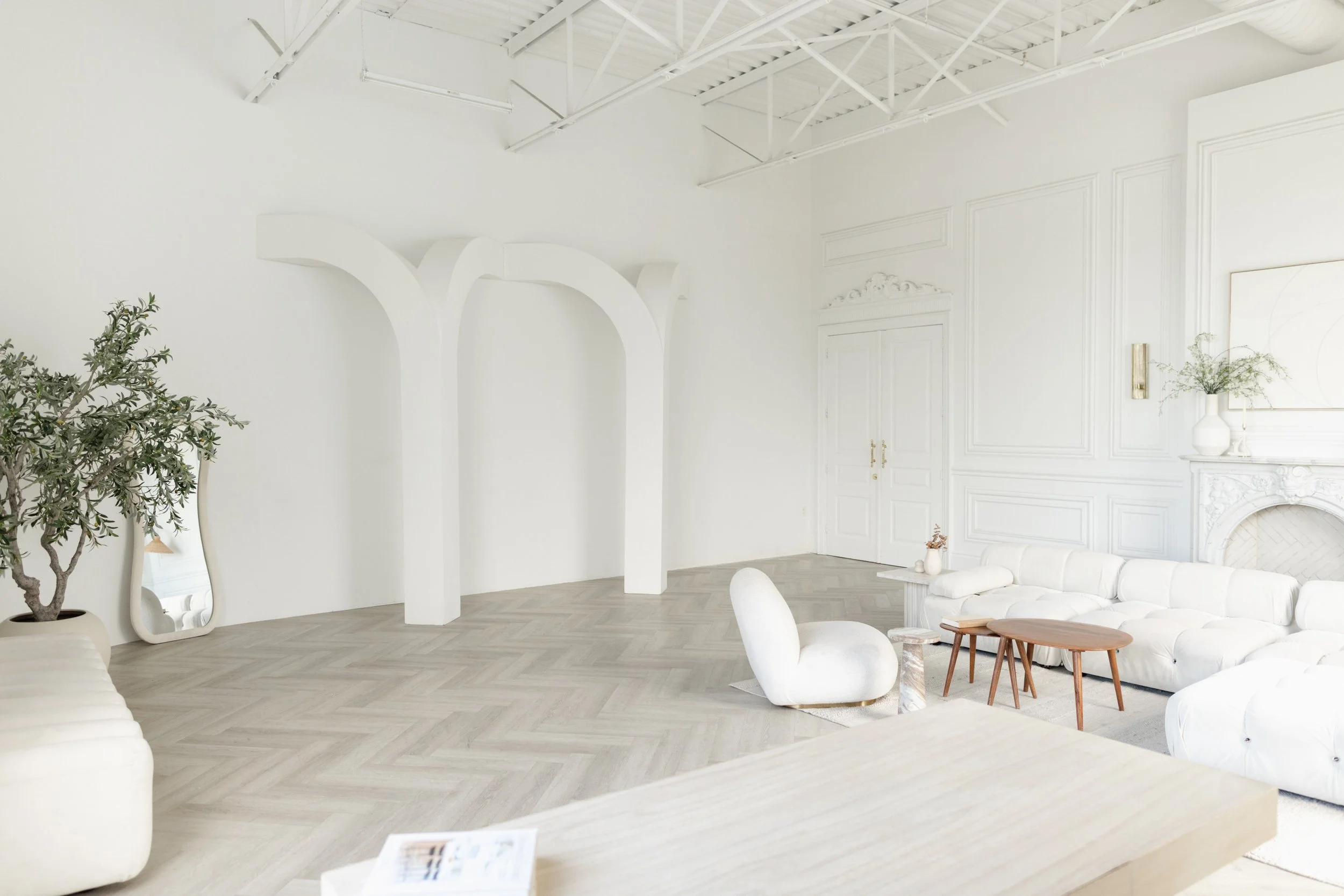 White living room with elegant decor, including a white sofa, coffee table, potted plant, and ornate wall molding, with a high ceiling and exposed beams.