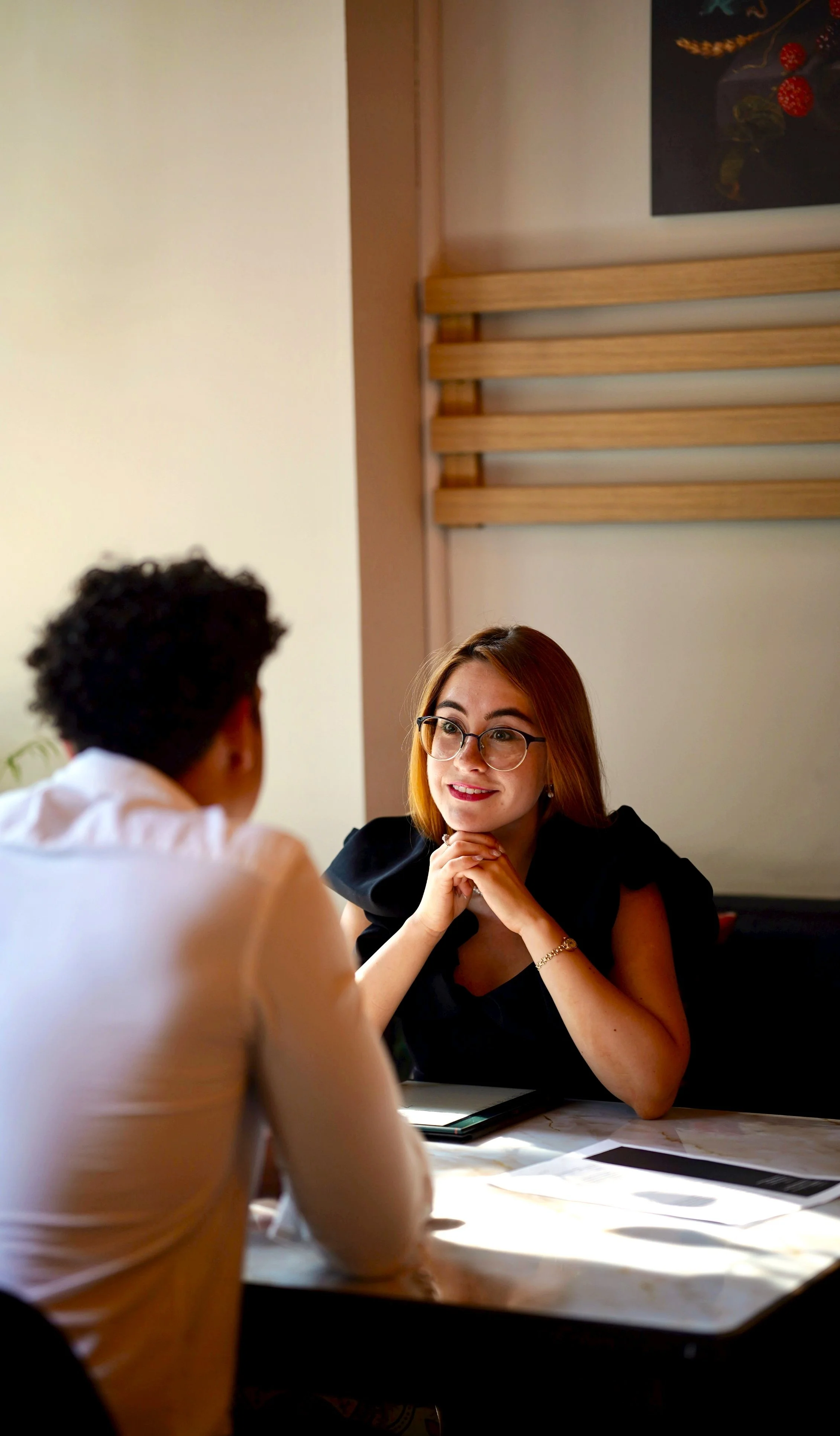 A woman with glasses and brown hair sits at a table, engaging in conversation with a person whose back is to the camera. The woman appears attentive with her hands clasped under her chin, smiling slightly. The setting looks like a casual indoor office or cafe with framed artwork and wooden wall accents.