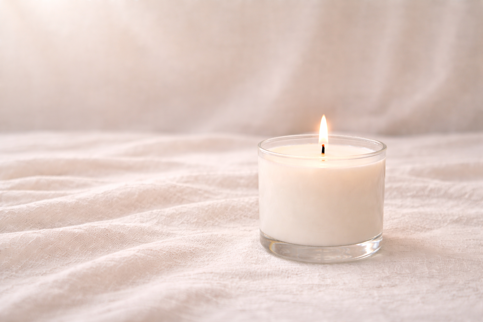 A lit white candle in a glass holder placed on a soft, textured surface with a neutral background.