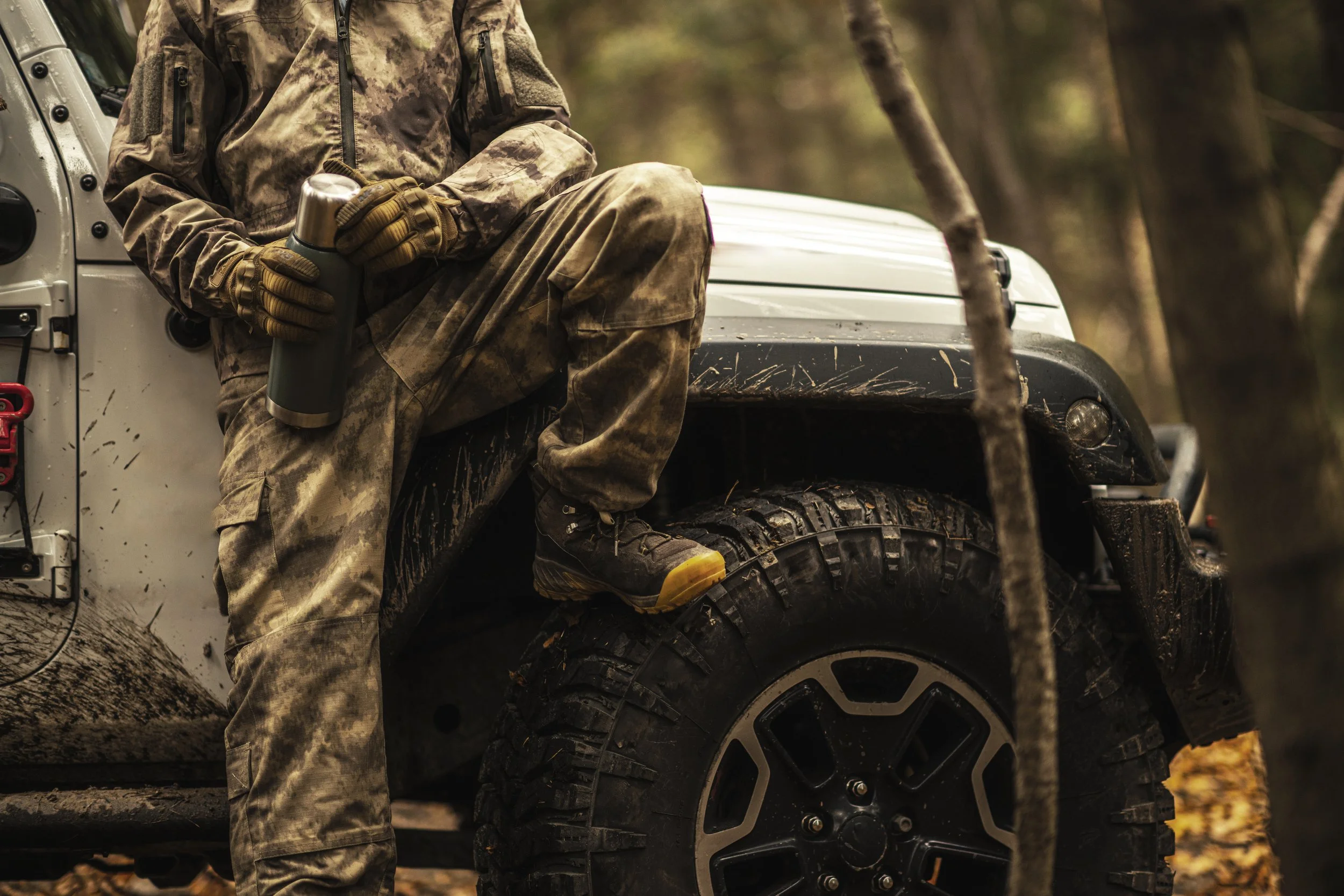 Person in camouflage holding a thermos, leaning against a muddy off-road vehicle in a forest.