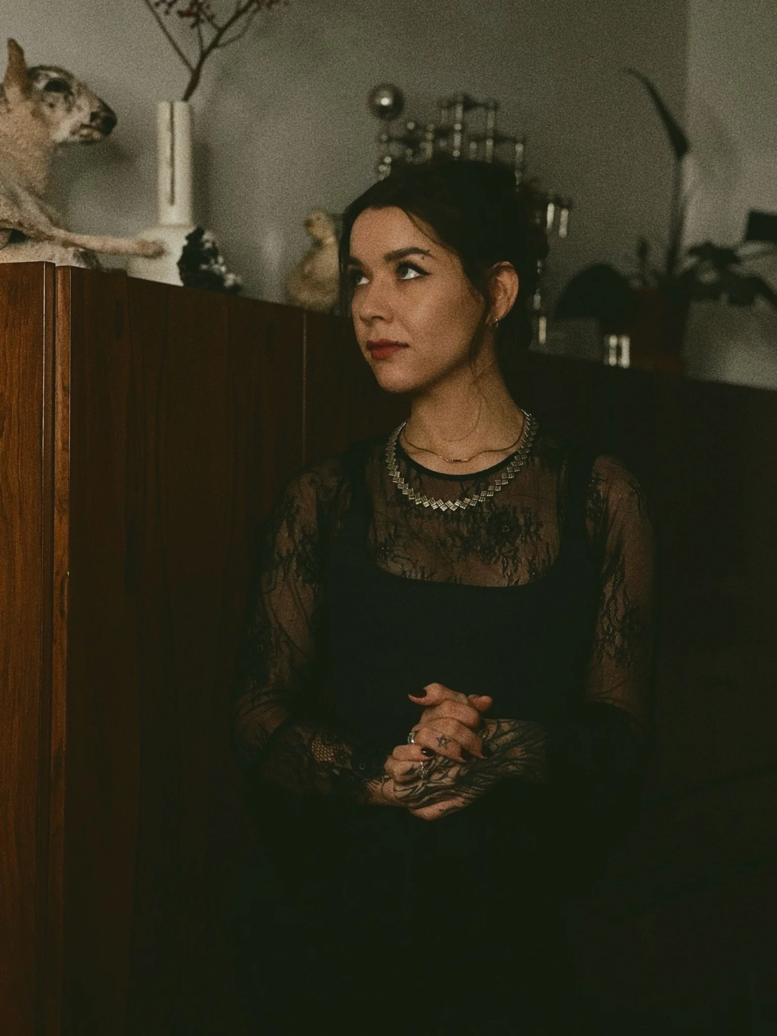 A woman with dark hair and tattoos on her hands, dressed in black lace, stands with her hands clasped in front of her, looking to the side. She is wearing layered necklaces and earrings, with a dark background featuring various decorative objects, including a mounted animal head, a vase, and sculptures in a dimly lit room.