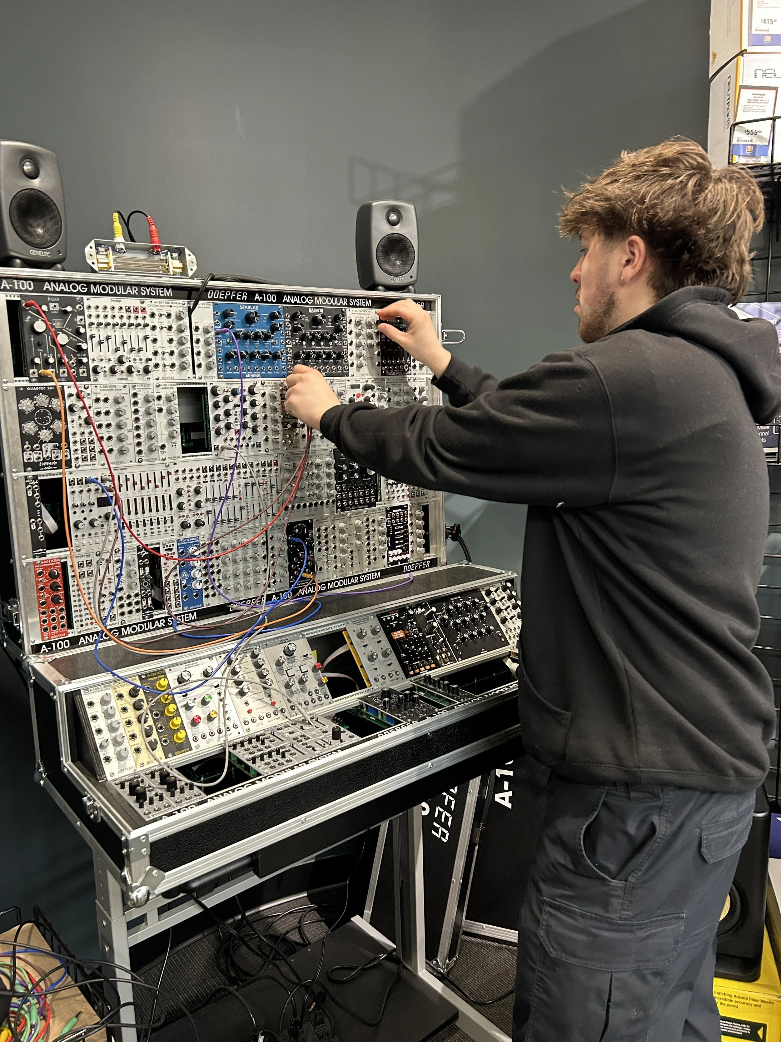 A person with brown hair wearing a black hoodie and black pants is working with an electronic modular synthesizer system on a stand. The setup includes various knobs, wires, and modules, with speakers on top and a wheel on the side.
