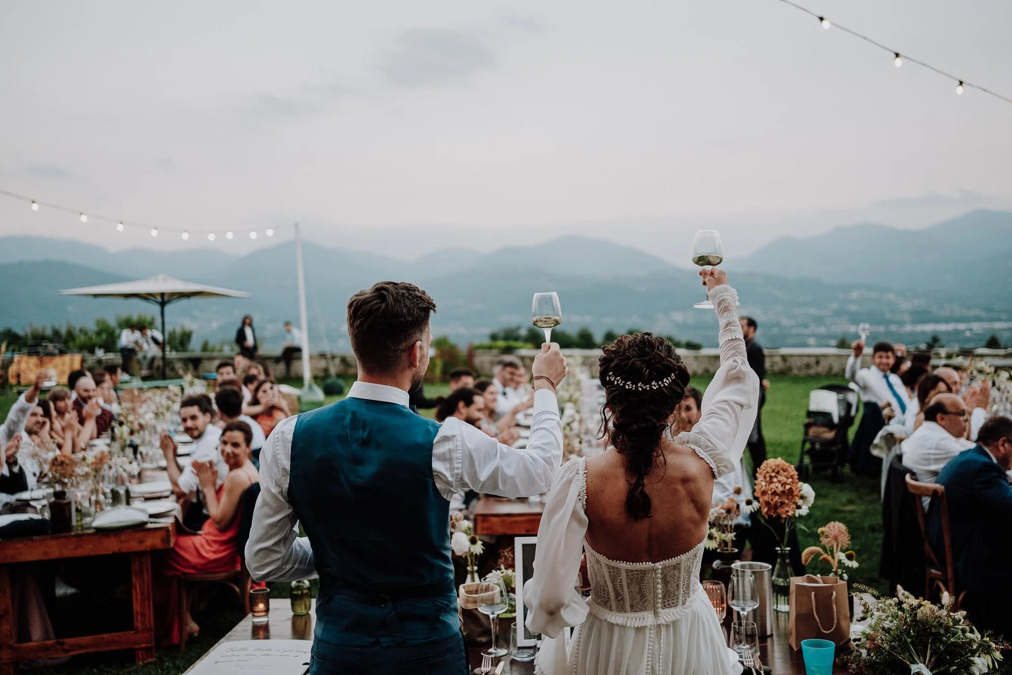 Sposi che brindano con bicchieri di vino durante un matrimonio all'aperto, con ospiti seduti a tavoli decorati con fiori, sullo sfondo di montagne e cielo nuvoloso.