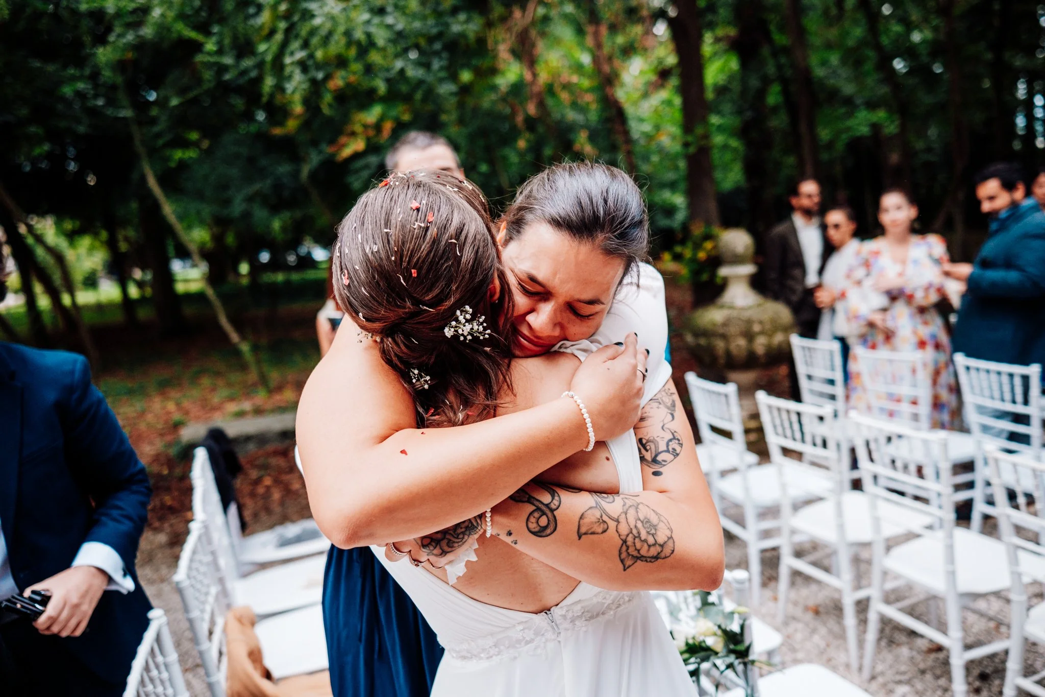 Due donne si abbracciano emozionate durante una cerimonia all'aperto, sedie bianche e persone in abiti eleganti sullo sfondo nel verde.