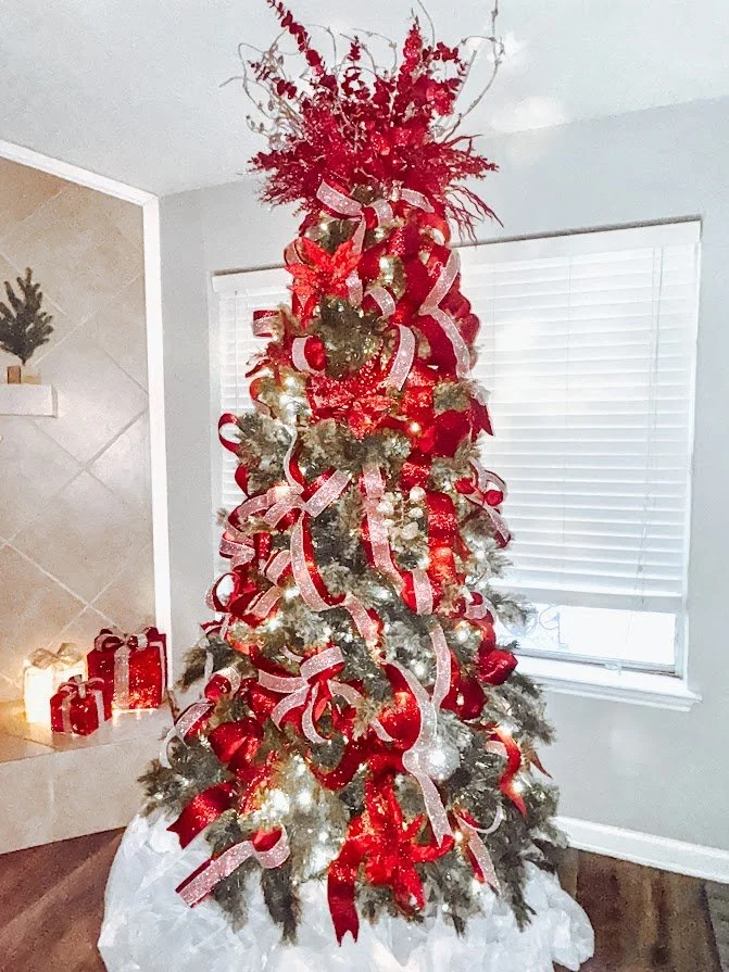 Decorated Christmas tree with red and white ribbons, ornaments, and a top arrangement of red branches, in a living room with a window and presents on a hearth.
