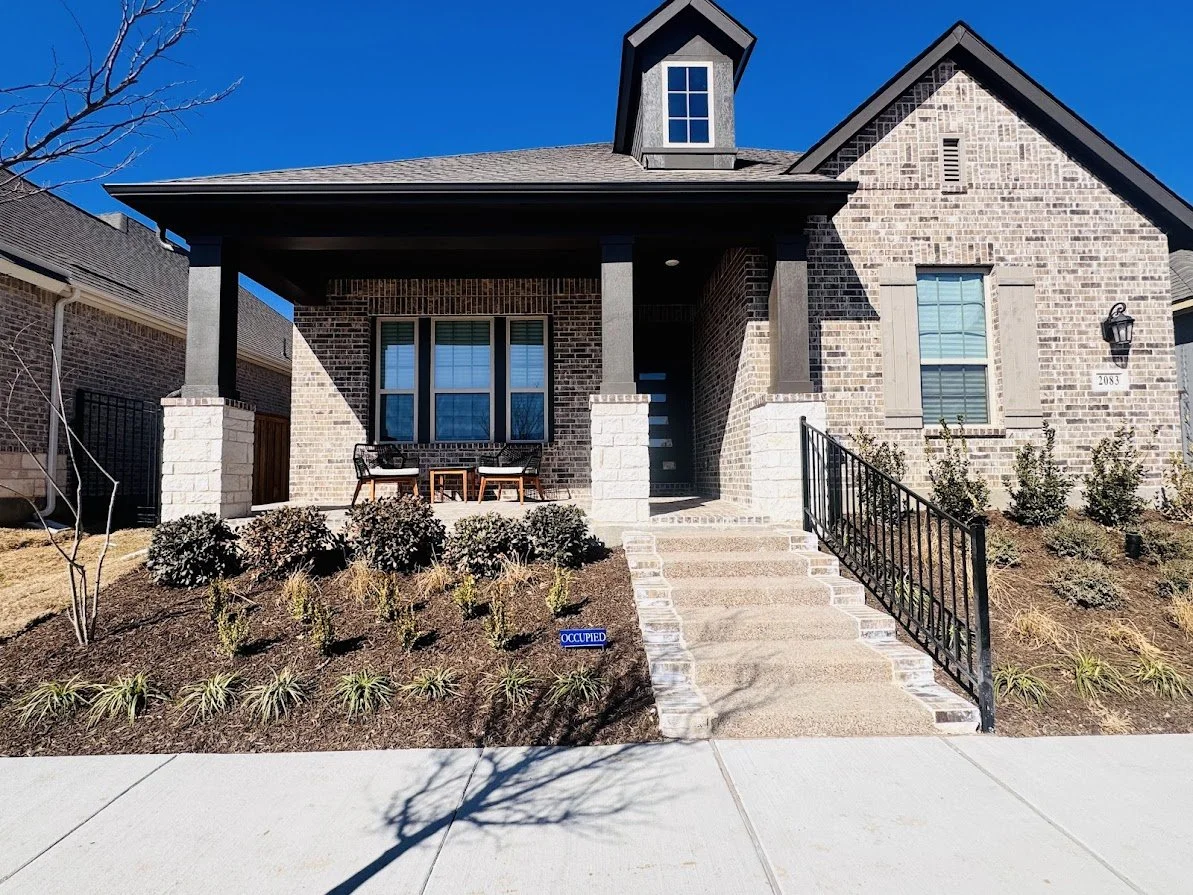 Front view of a brick suburban house with a porch, stairs, and landscaped yard under a clear blue sky.