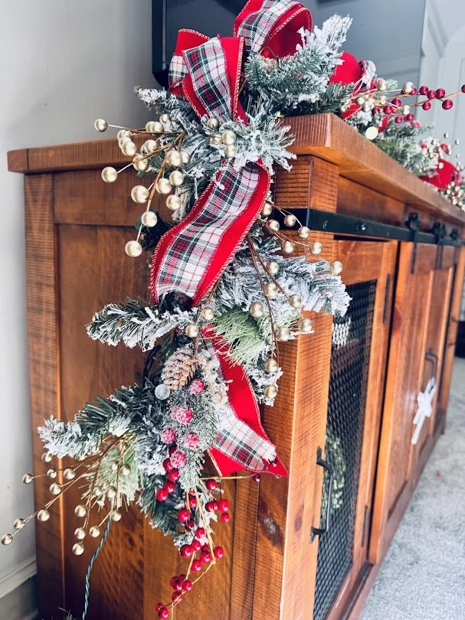 Christmas garland with red plaid ribbons, faux snow, red berries, pinecones, and gold beads decorating a wooden cabinet