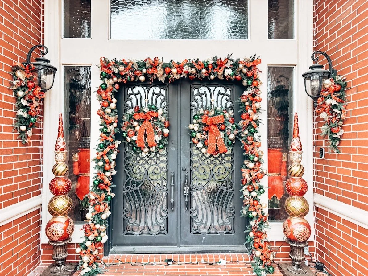 Decorated front door with Christmas garland and ornaments, flanked by wall-mounted lanterns and large decorative vases.