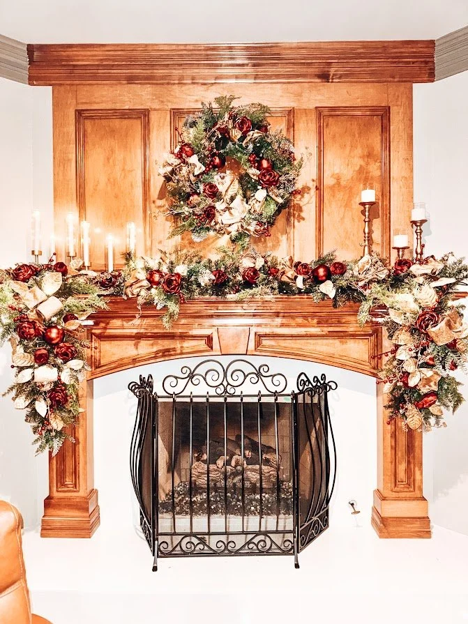 Decorated wooden fireplace mantel with Christmas wreath, garland, red and gold ornaments, candle holders, and a fireplace with logs.
