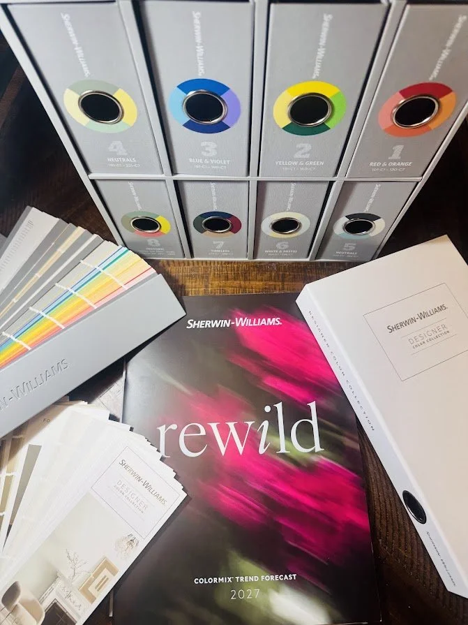Color swatches, a Sherwin-Williams color forecast book titled 'Rewild' for 2027, and several sample cards for interior paint colors on a wooden surface.
