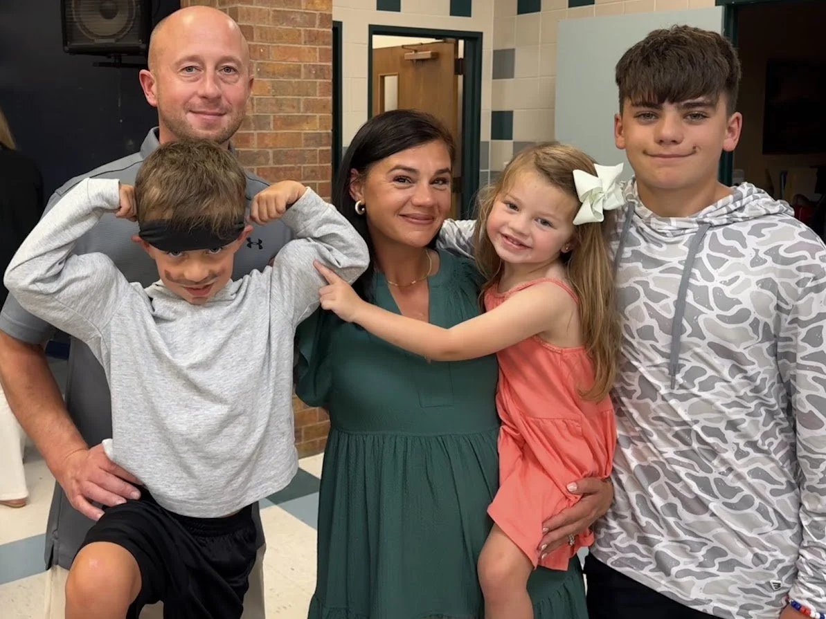 A group of five people, including two adult men and women and three children, smiling and posing together in a school or community center setting.