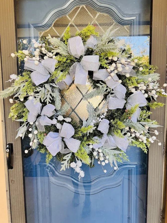 A decorative holiday wreath with green foliage, white berries, and large gray bows hanging on a blue front door.