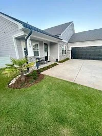 Front yard of a house with a well-maintained lawn, a garden bed with a small palm plant, and a driveway leading to a two-car garage.
