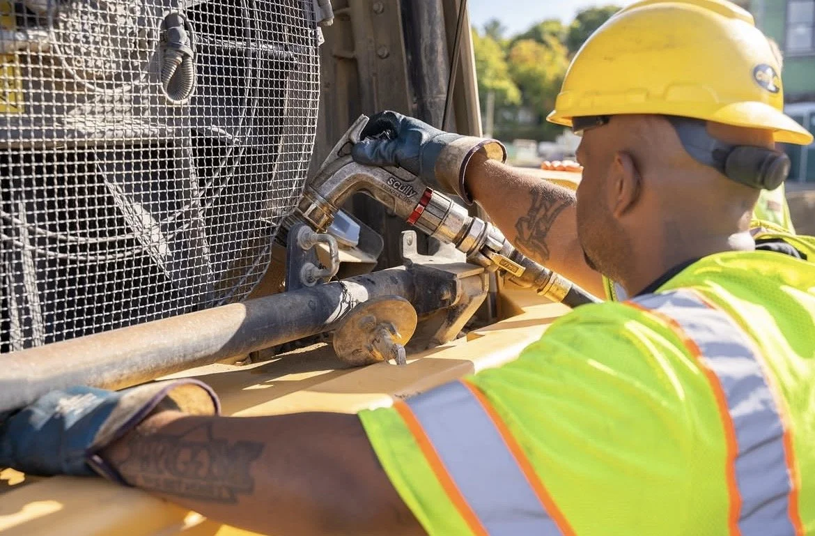 A construction worker wearing a yellow hard hat, black gloves, and a neon yellow safety vest using a wrench on heavy machinery at a construction site.