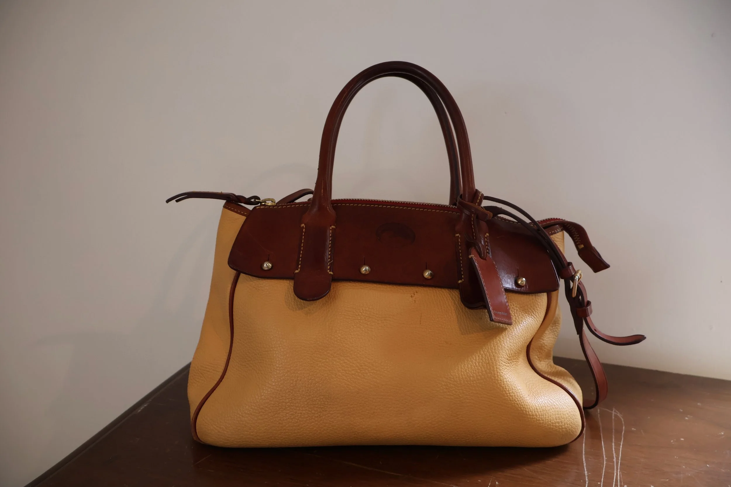 A beige leather handbag with brown leather handles and a flap, placed on a wooden surface against a plain wall.