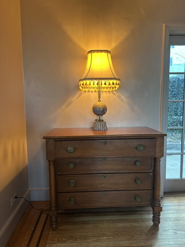 A wooden chest of drawers with a lamp on top, positioned in front of a wall near a glass door.