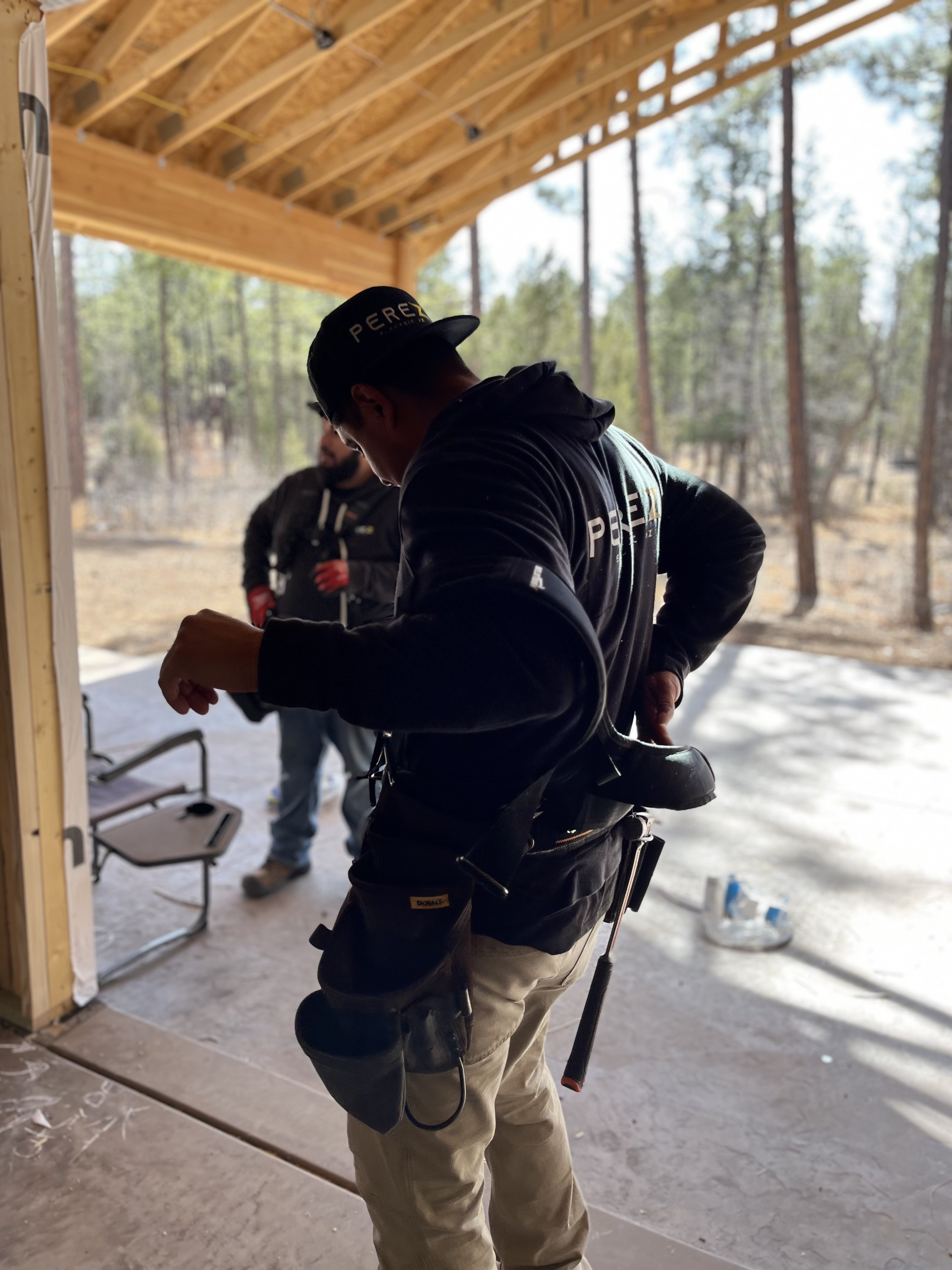 Construction worker wearing tool belt and cap adjusting gear on worksite with wooden roof framework and forest view.
