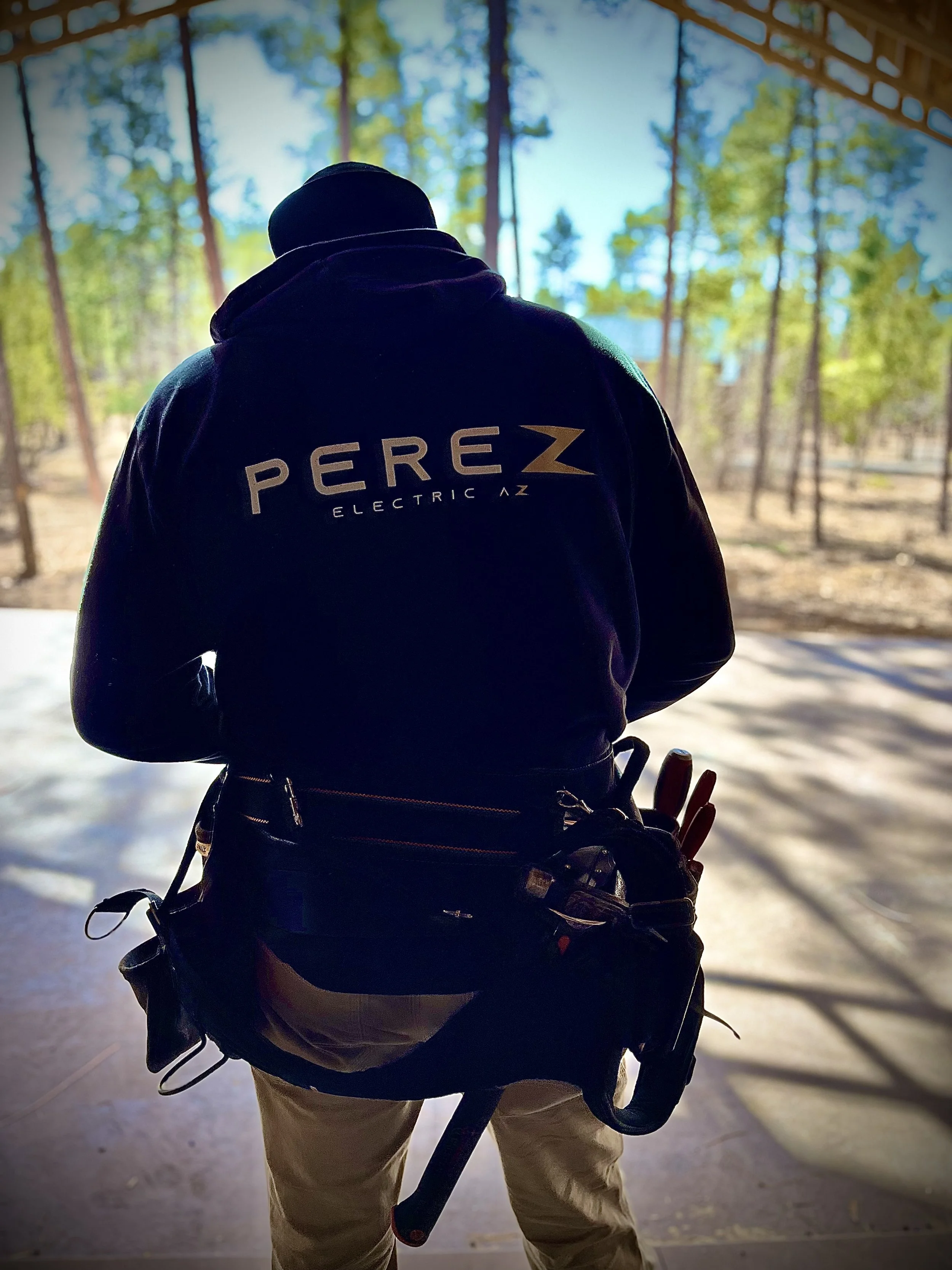 Back view of a person wearing a "Perez Electric AZ" hoodie and a tool belt, standing outdoors with trees in the background.