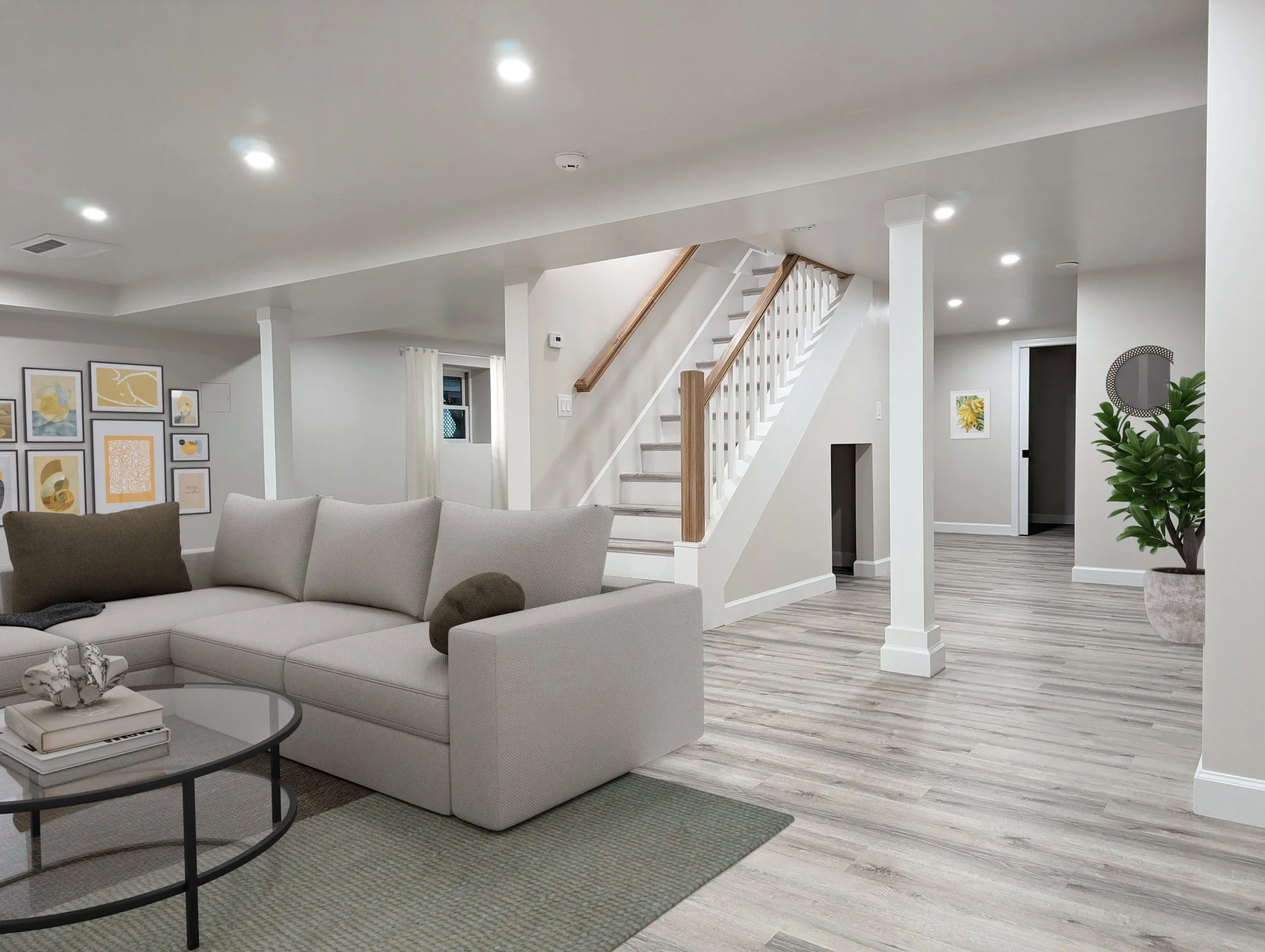 A modern living room with a white sectional sofa, black coffee table with books and decorative objects, artwork on the gray walls, a staircase with wooden handrails, and a plant in the corner.