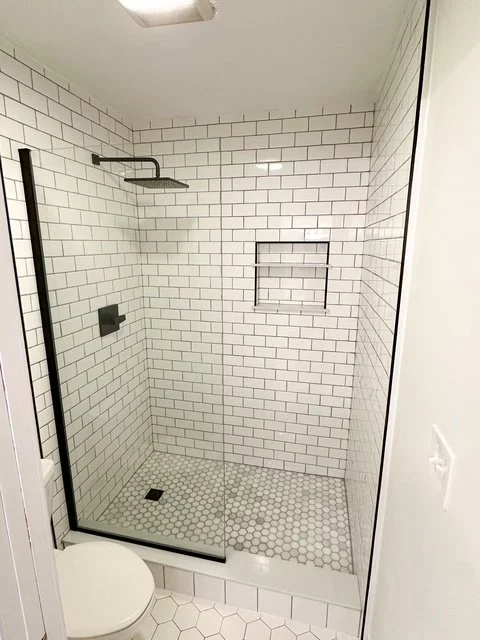 Bathroom with a glass shower enclosure, white subway tiles, a showerhead, a built-in shelf, and a toilet.
