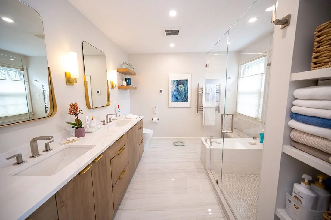 Bright modern bathroom with double vanity sinks, large mirrors, a decorative plant, artwork, and a walk-in shower with glass enclosure, along with a bathtub, towel racks, and neatly folded towels.