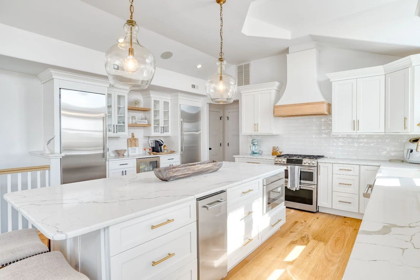 Modern white kitchen with marble countertops, gold accents, hardwood flooring, and pendant lighting.