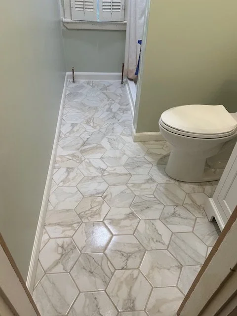 A small bathroom with hexagonal marble-patterned tile flooring, a toilet on the right, a light green wall, a window with shutters at the back, and a shower curtain partially visible on the right.