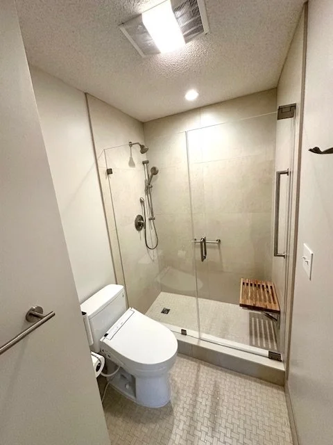 Modern bathroom with a glass-enclosed walk-in shower, a white toilet, and a small wooden bench.