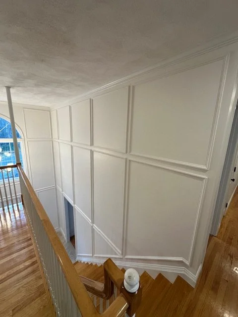Interior view of a staircase landing with white paneled walls, a wooden handrail, hardwood flooring, and a large arched window in a residential home.
