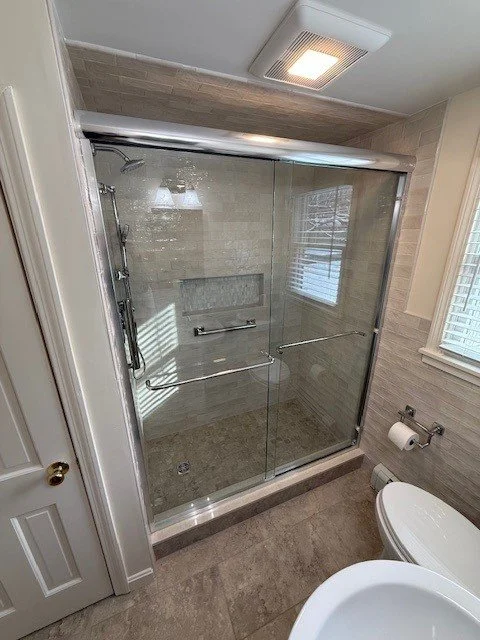 Bathroom with glass shower enclosure, beige tiles, a window with blinds, and a toilet.