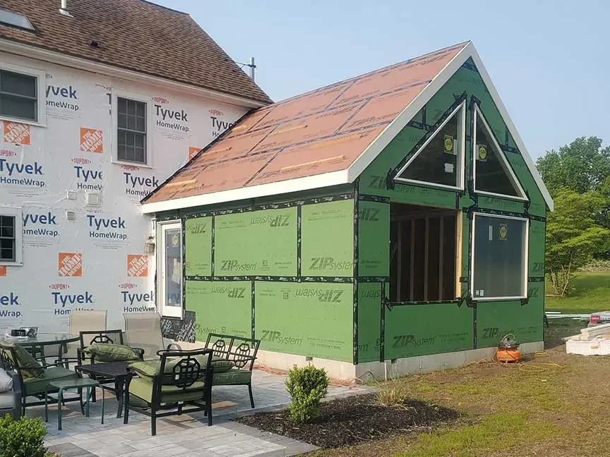 A small house under construction with green sheathing, a pitched roof, and a triangular window in the front. A patio area with outdoor furniture is in the foreground.
