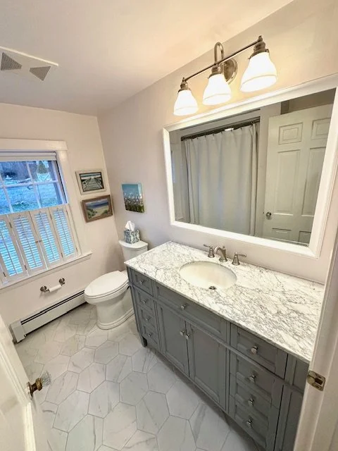 Bathroom with a marble countertop vanity, a mirror, a light fixture with three lights, a toilet, a window with blinds, and artwork on the wall.