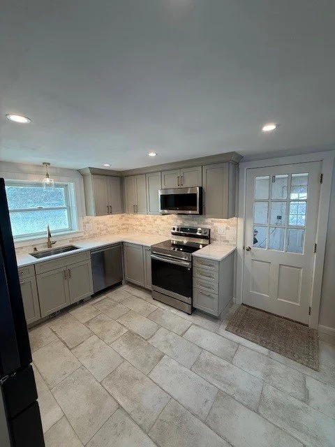Empty kitchen with gray cabinets, stainless steel stove, white tile backsplash, window above sink, and a door leading outside.