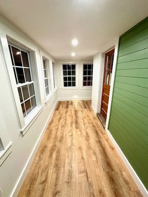 Bright enclosed porch with multiple large windows, wood flooring, and a green paneled wall with a brown front door.