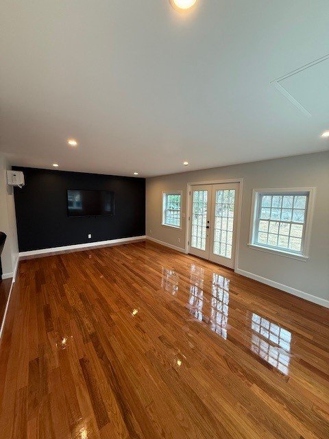 Empty living room with hardwood floors, white walls, a black accent wall with a mounted TV, multiple windows, and a door leading outside.