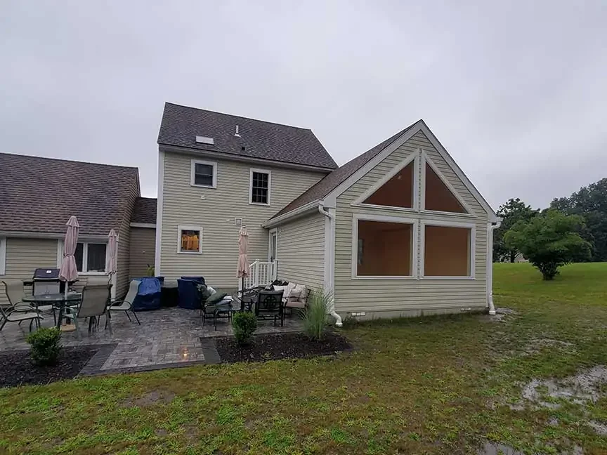 Backyard of a house with patio furniture, umbrellas, a grill, and screened-in porch under construction on a cloudy day.