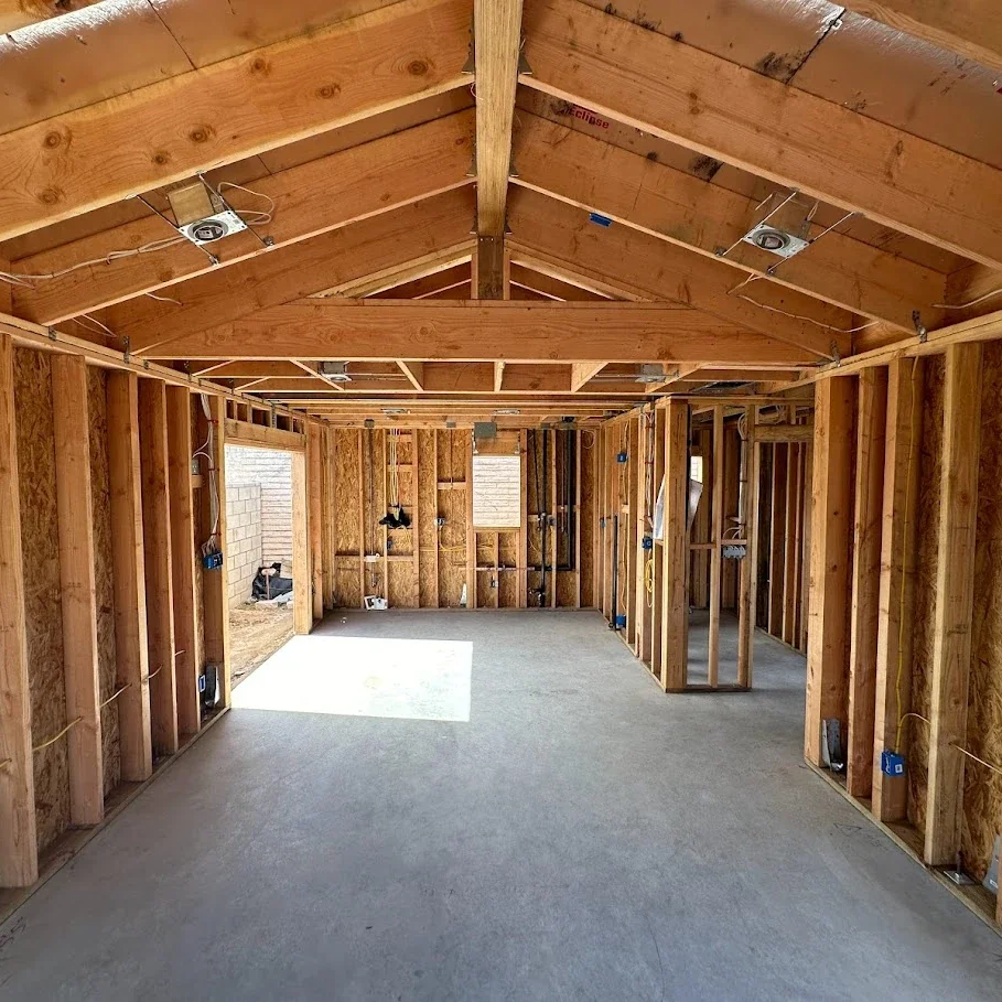 Interior of a house under construction with exposed wooden framing, electrical wiring, and a concrete floor, with windows on each side.