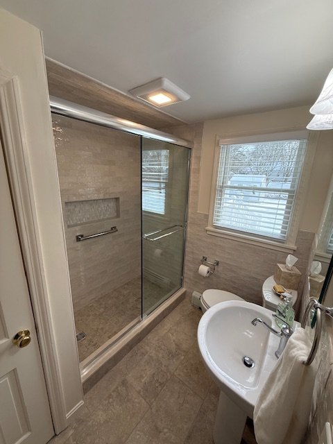 Small bathroom with a glass shower enclosure, a window with blinds, a white toilet, and a pedestal sink with a towel and toiletries.