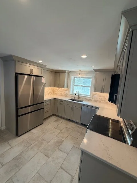 Modern kitchen with stainless steel refrigerator, sink under a window, light gray cabinets, marble countertops, and a white tile backsplash.