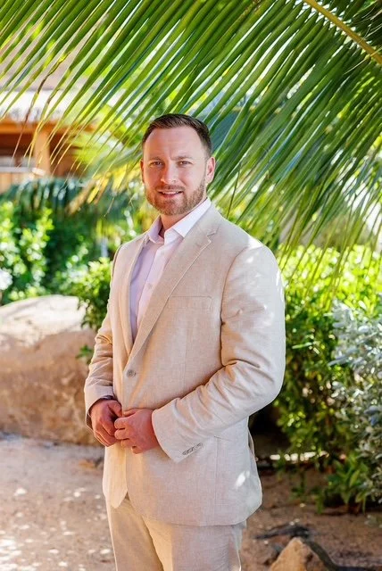 A man in a beige suit standing outdoors surrounded by lush greenery and palm leaves.