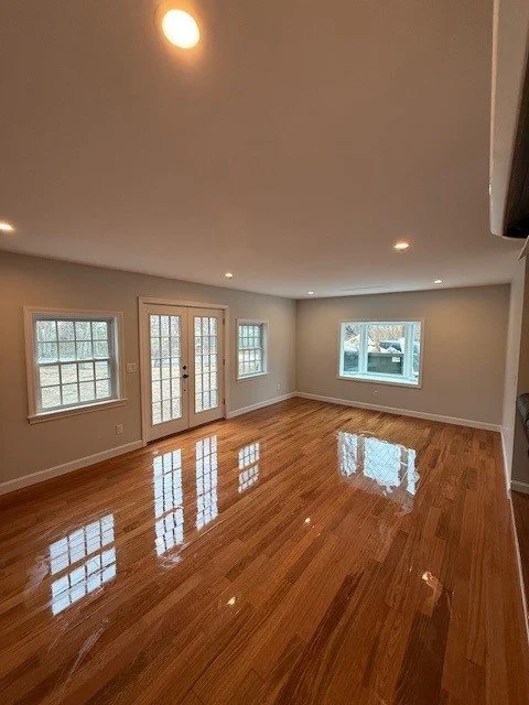 Empty living room with hardwood floors, windows, and glass doors leading outside, under a ceiling with recessed lighting.