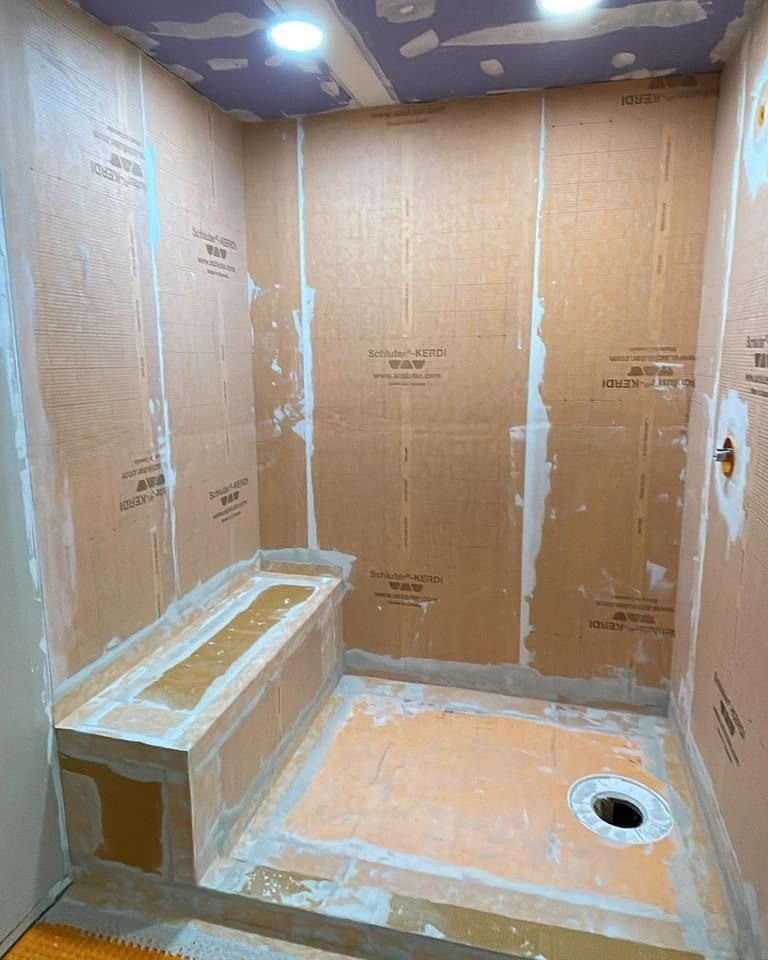 A small bathroom under construction, with unfinished walls covered with insulating and moisture barrier panels, a floor drain, and a step with a tub outline.