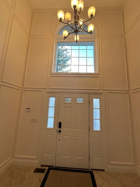 Entryway with a white front door, two narrow windows on sides, large window above, chandelier hanging from ceiling, and a doormat on tile floor.