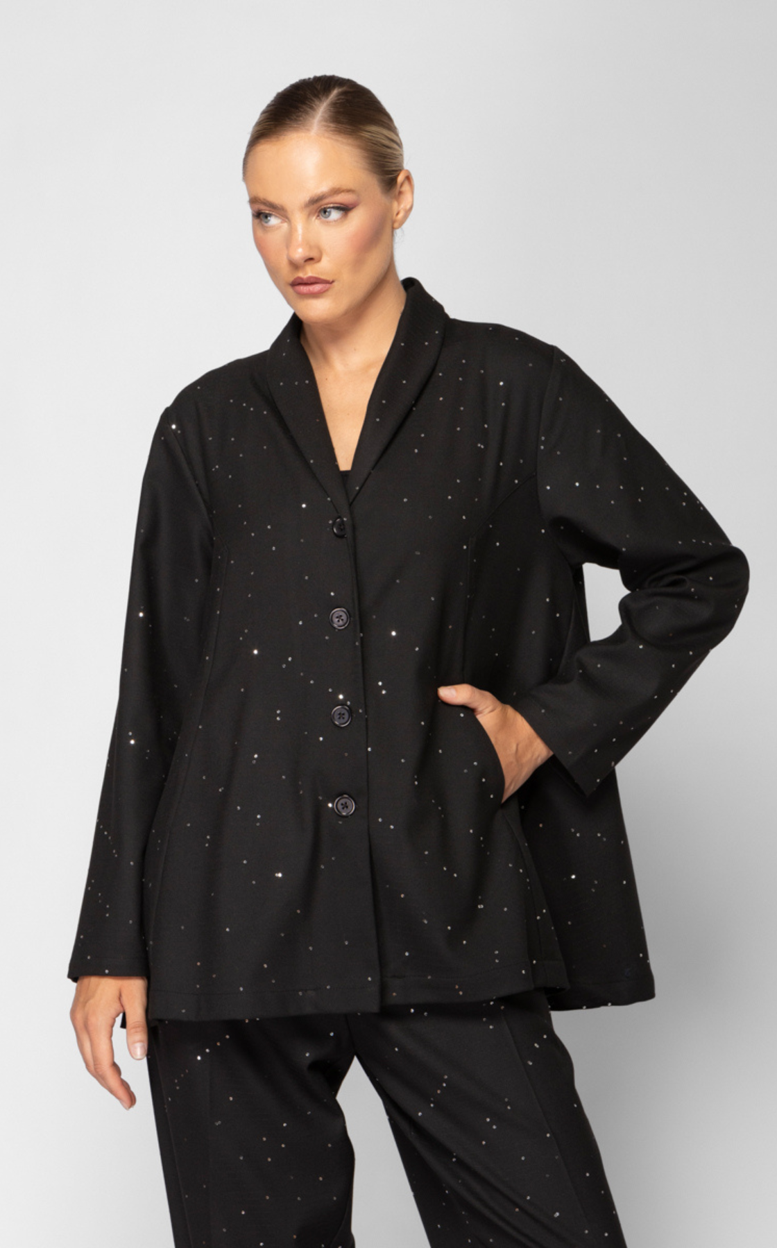 A woman wearing a black button-up jacket and matching pants with silver speckles, posing with one hand in her pocket against a plain gray background.