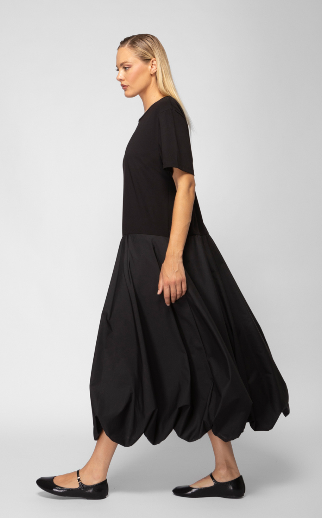 A woman walking with a long black puffed skirt, black top, black flats, and blonde hair against a plain background.