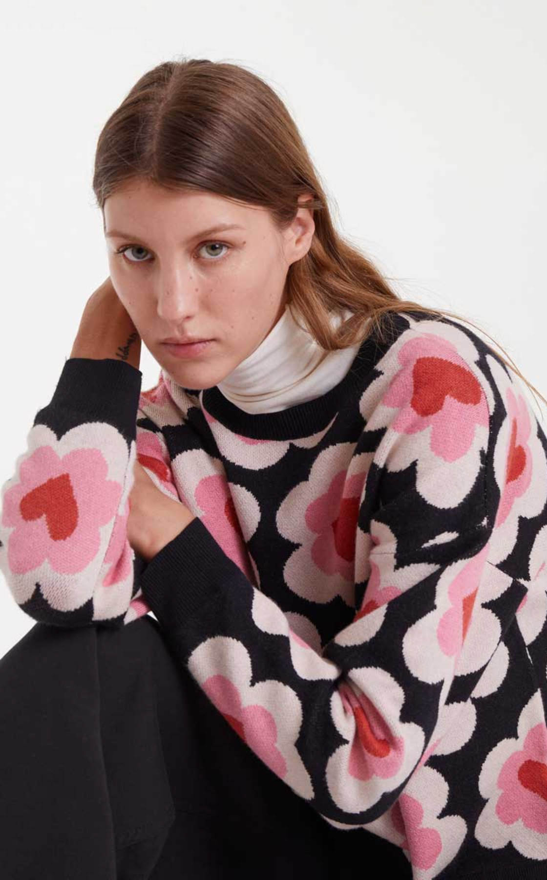A young woman with red hair wearing a black sweater with pink and red floral patterns and a white turtleneck underneath.