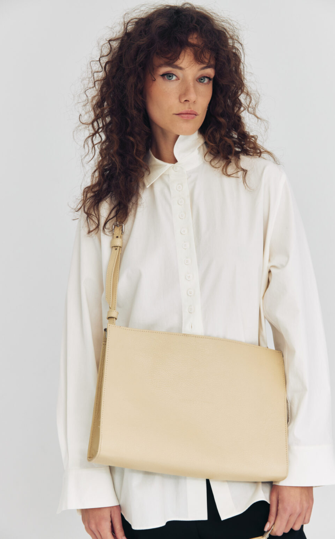 A woman with curly brown hair and blue eyes wearing a white button-up shirt, holding a beige handbag with a strap.