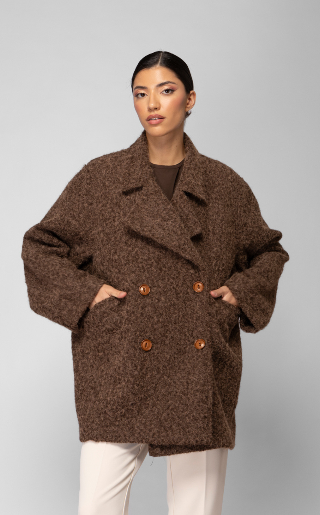 Woman with dark hair in a sleek ponytail wearing a double-breasted brown wool coat and light beige pants against a plain gray background.