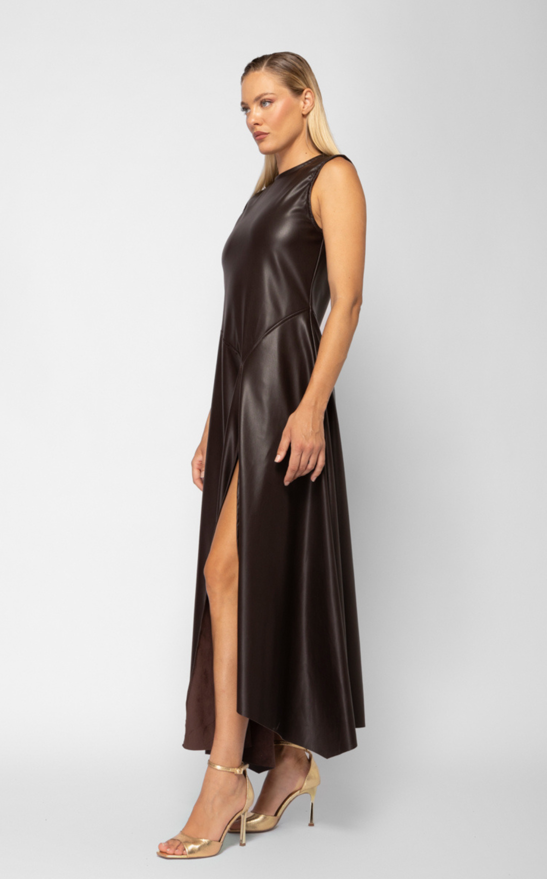 A woman wearing a sleeveless, dark brown, leather-like gown with a high slit, gold high heels, and standing against a plain white background.