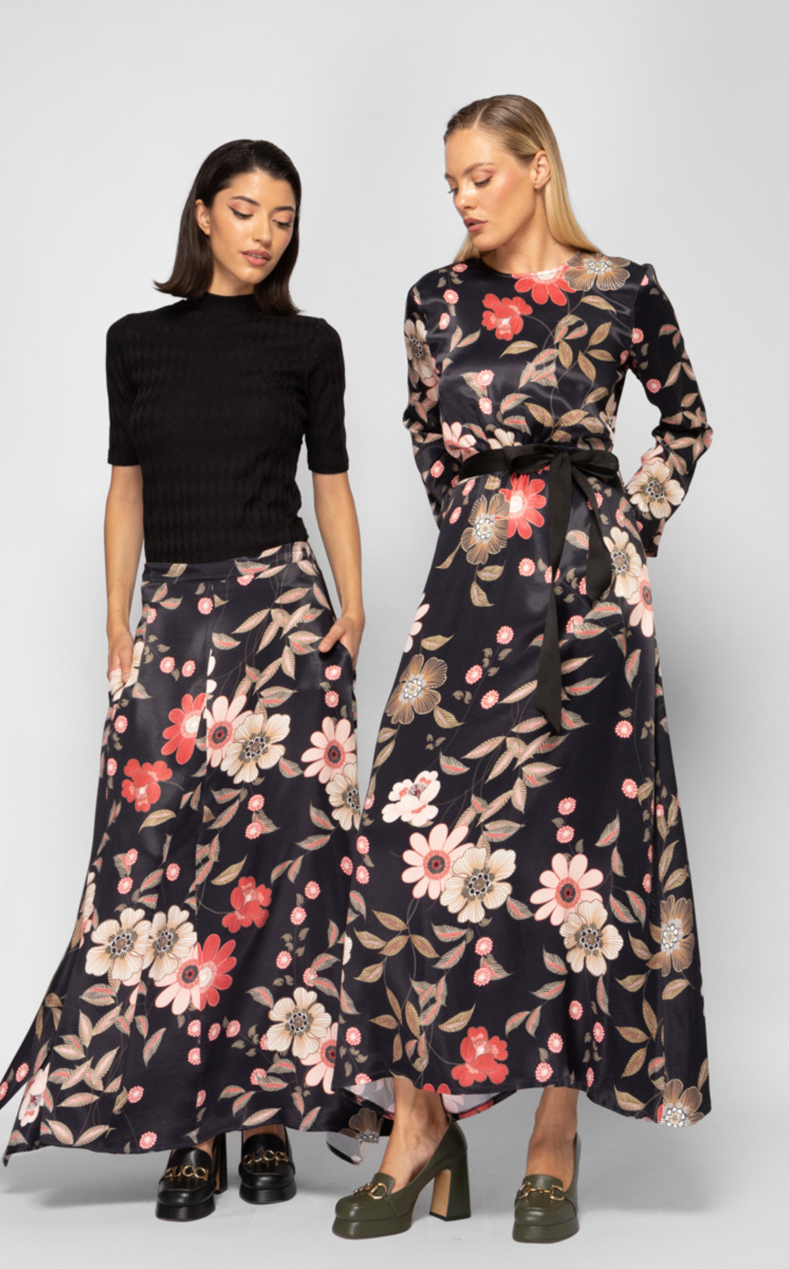 Two women wearing matching floral long skirts, one in black top and the other in a floral dress, standing against a plain background.
